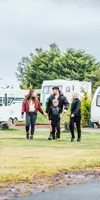 A family of two parents and two children wearing coats and hats walking on a green field in front of touring vans and motorhomes 
