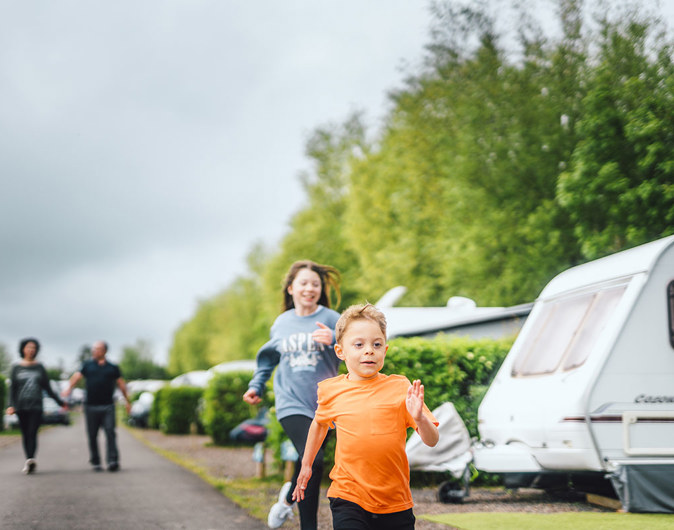 Two children, a boy and a girl, are running past touring vans next to trees and two adults walk in the background 