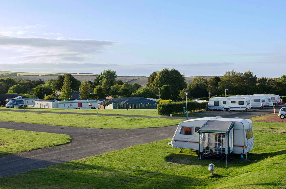 A high angle image of touring vans and motorhomes parked on touring pitches in a grassy field at a holiday park on a sunny day 