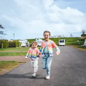 Two little girls in matching colourful cardigans holding hands and running next to touring caravans on pitches 
