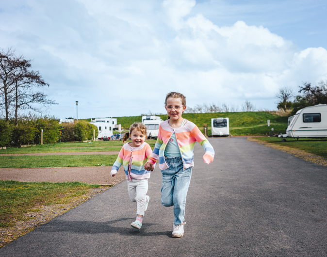 Two little girls in matching colourful cardigans holding hands and running next to touring caravans on pitches 