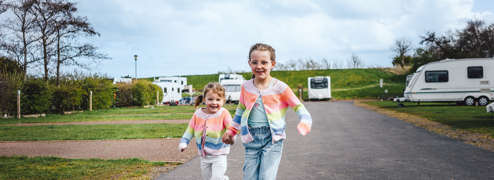Two little girls in matching colourful cardigans holding hands and running next to touring caravans on pitches