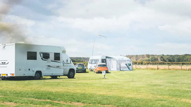 Two touring vans and cars parked on touring pitches at a UK holiday park with green grass and a blue sky above 