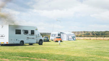 Two touring vans and cars parked on touring pitches at a UK holiday park with green grass and a blue sky above 