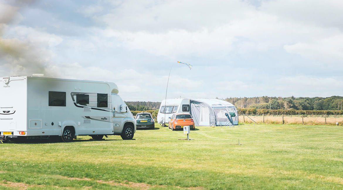 Two touring vans and cars parked on touring pitches at a UK holiday park with green grass and a blue sky above
