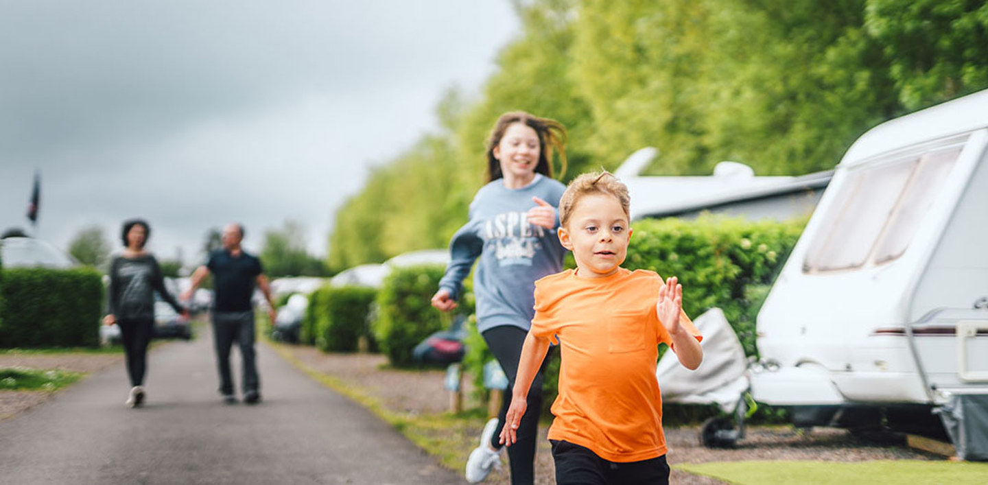 A young boy and young girl running next to touring vans on a holiday park, their parents are walking behind them